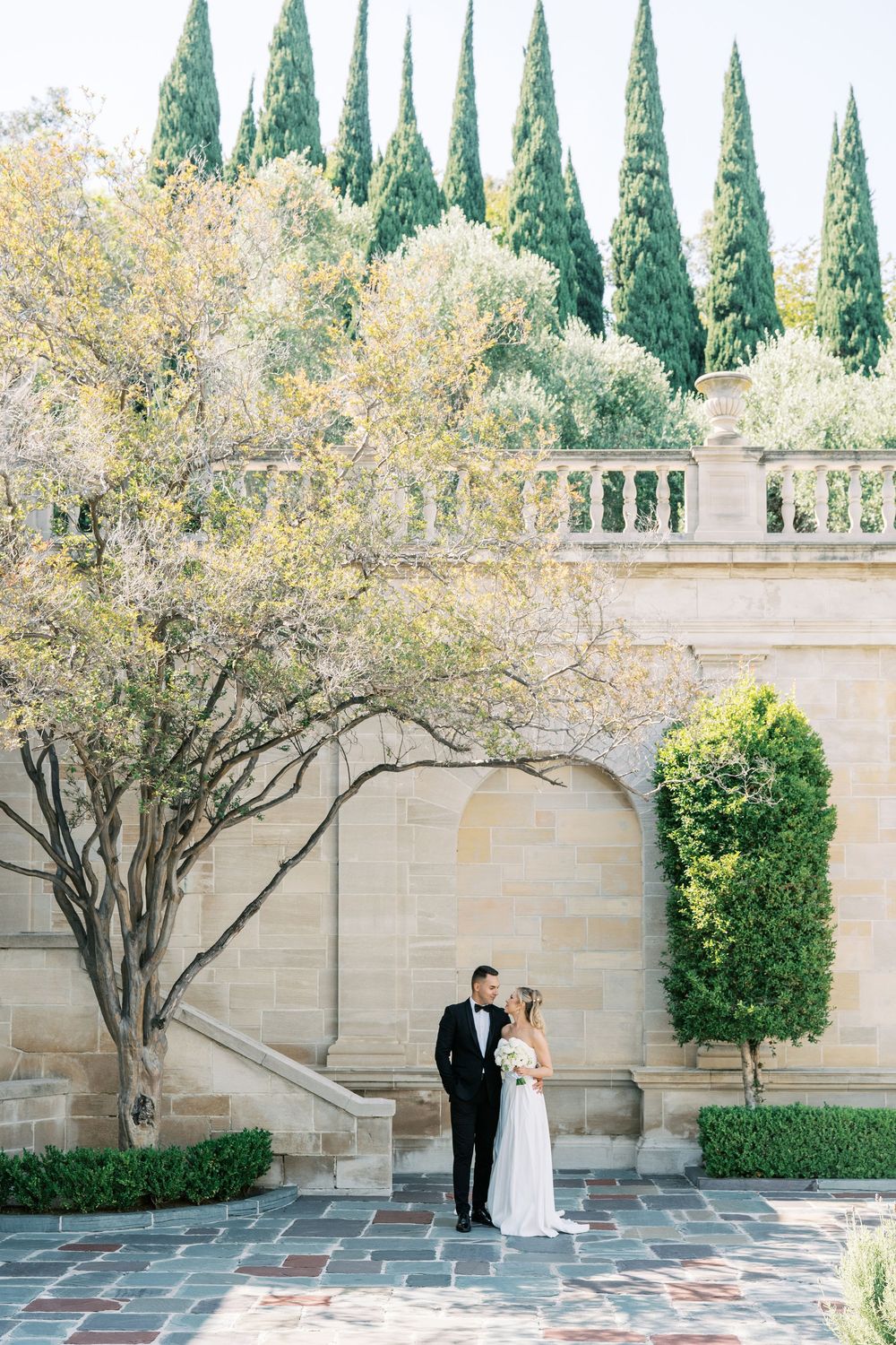 Couple shares romantic moment against limestone wall with cypress trees and climbing vines in elegant garden setting.