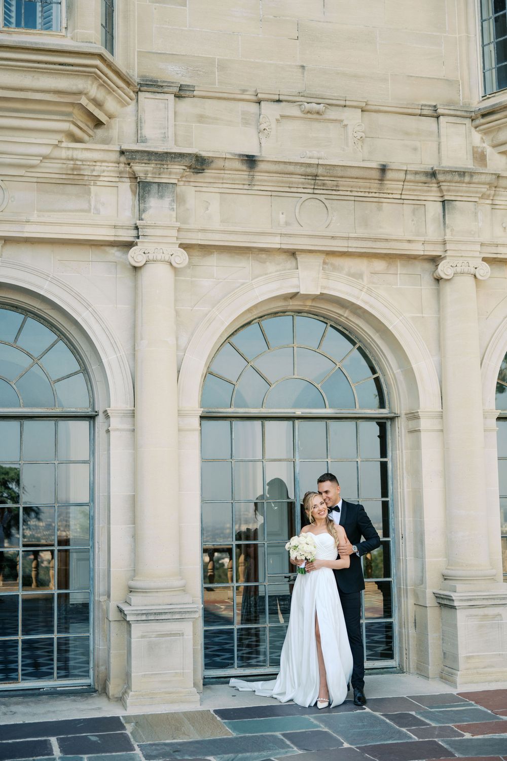 Elegant couple in wedding attire stands before grand arched windows at ornate stone building.