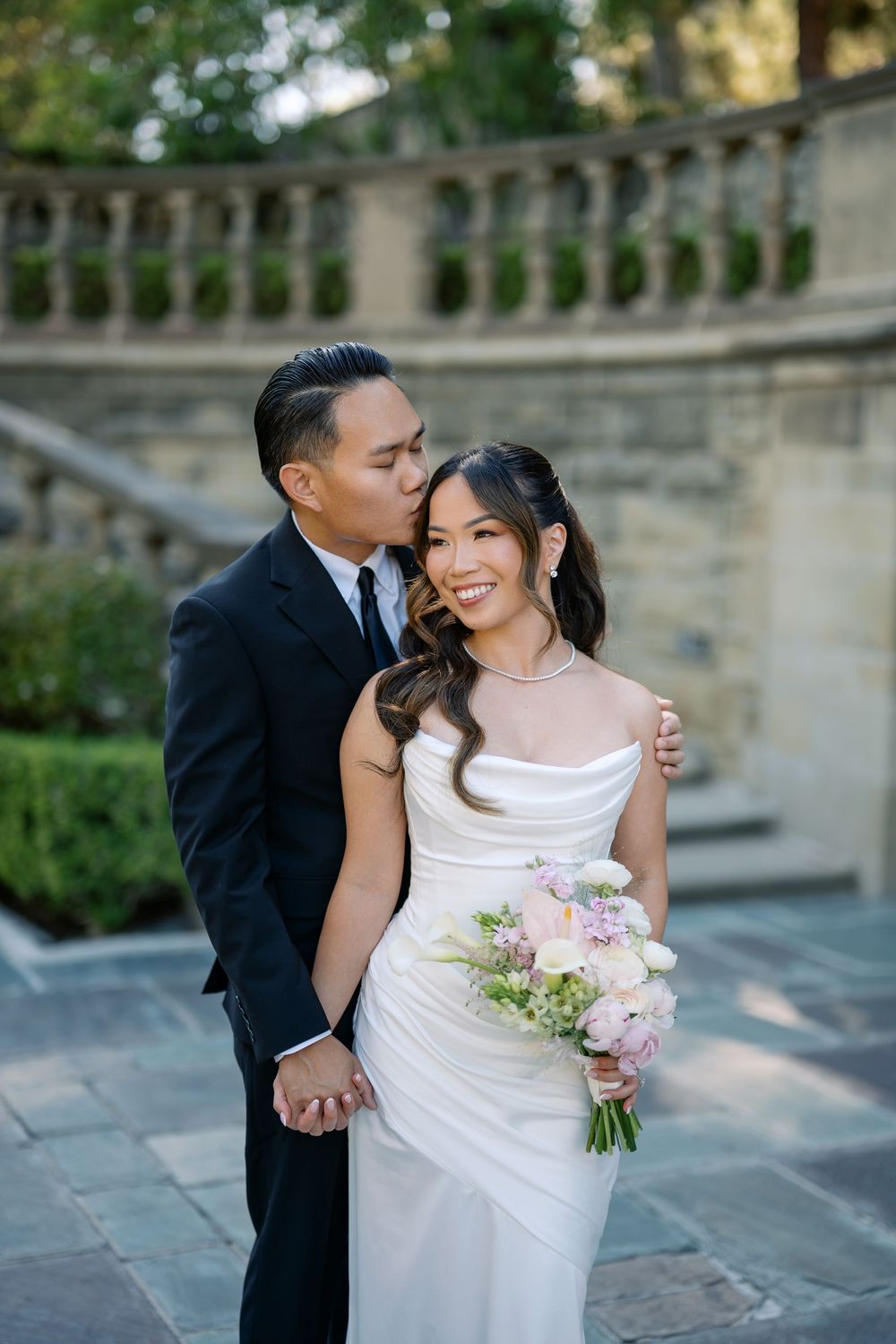 Groom in black suit kisses bride wearing white satin gown holding pink floral bouquet at elegant stone venue.