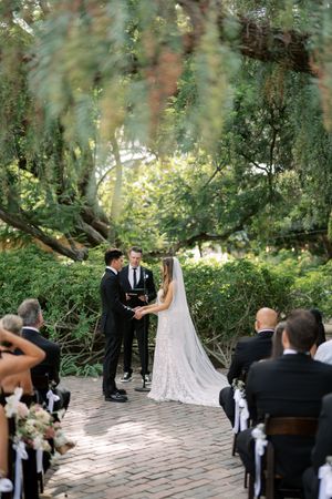 Bride and groom exchange vows outdoors under lush green trees with seated wedding guests in attendance.