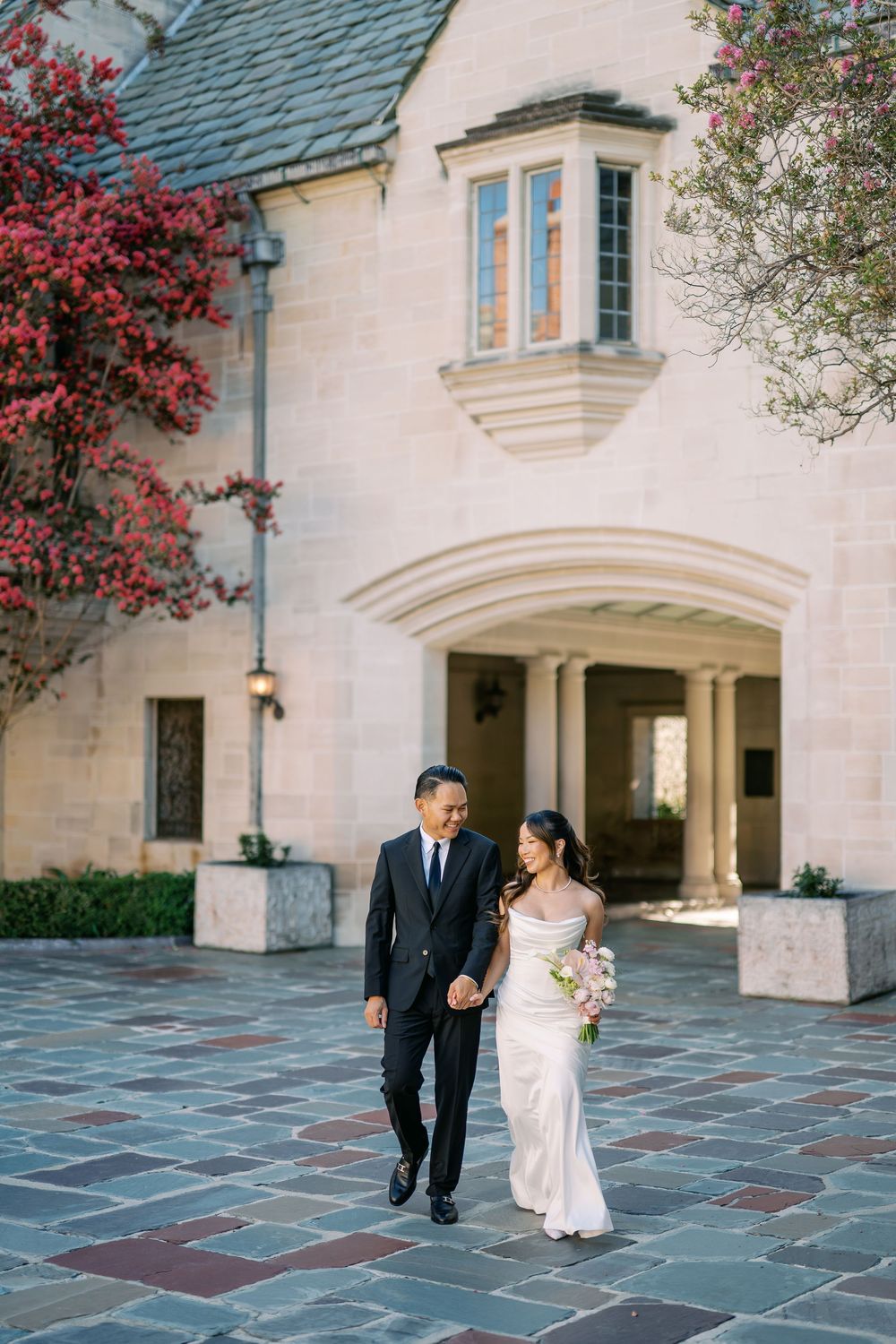 Asian bride and groom walk hand-in-hand outside an elegant stone manor with blooming red bougainvillea.