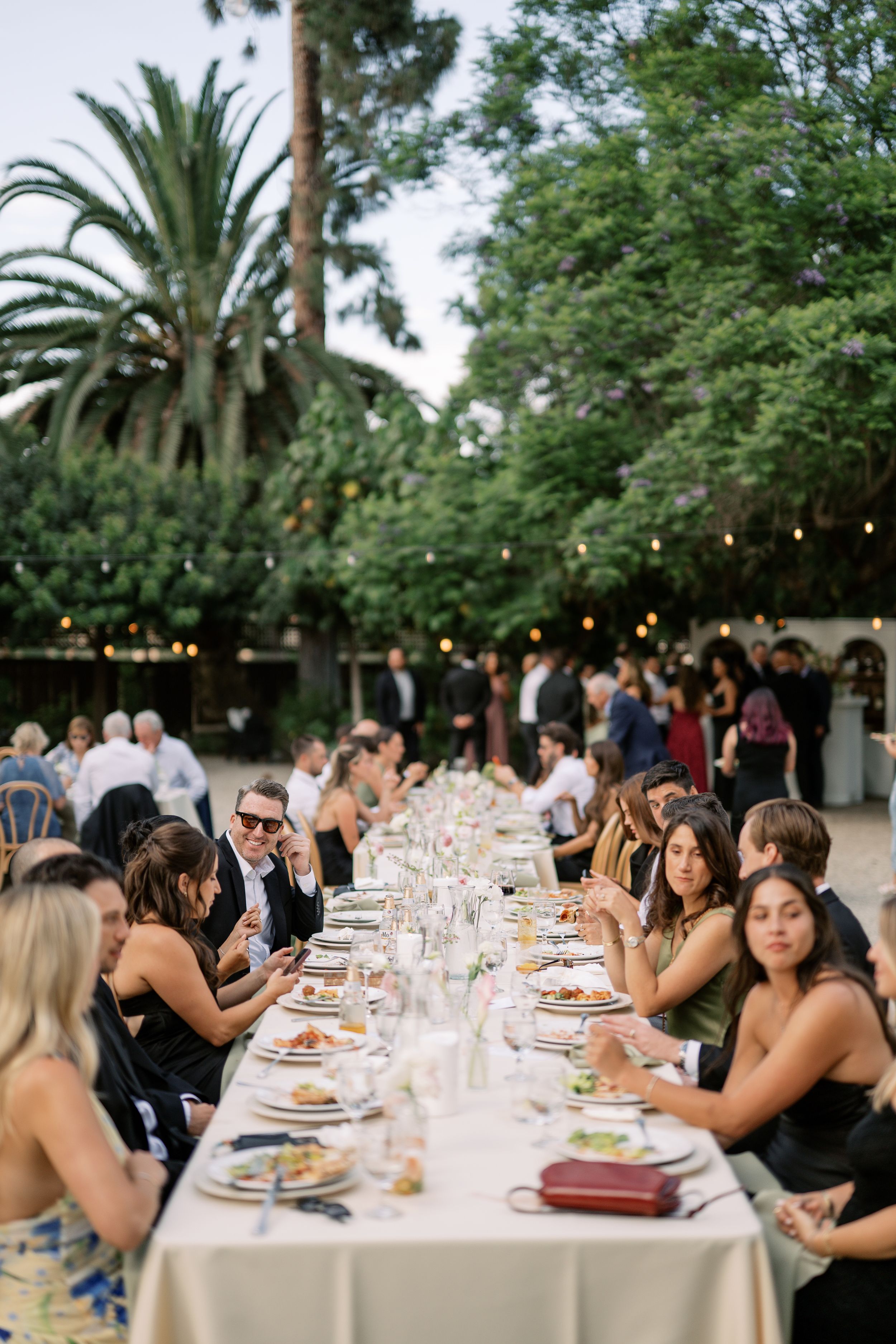 Guests dining at a long outdoor wedding reception table surrounded by lush trees and string lights at dusk.