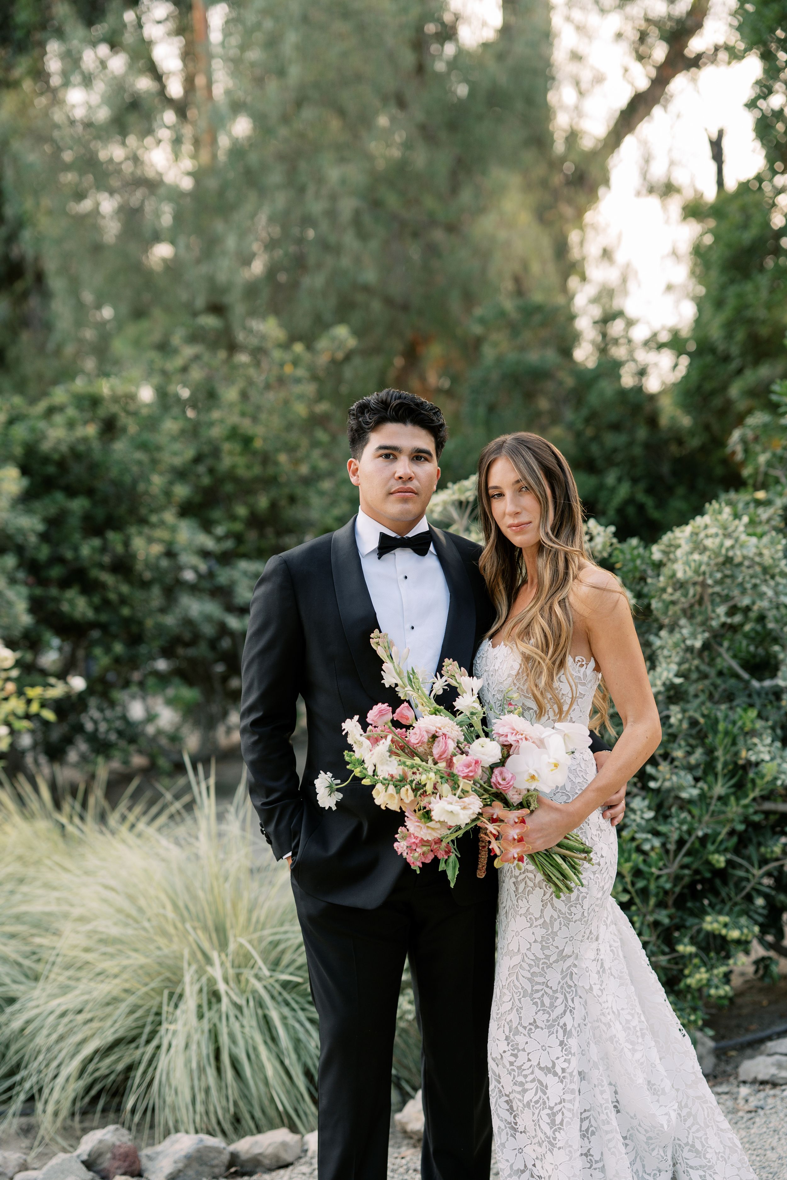 Elegant bride and groom posing outdoors in lush garden, wearing lace wedding gown and black tuxedo with pink floral bouquet.