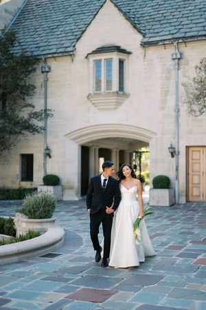 A romantic wedding couple walking along a curved stone courtyard in front of a traditional French-style building.