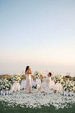 Man proposes on one knee surrounded by white flowers, candles, and rose petals with ocean view at sunset.