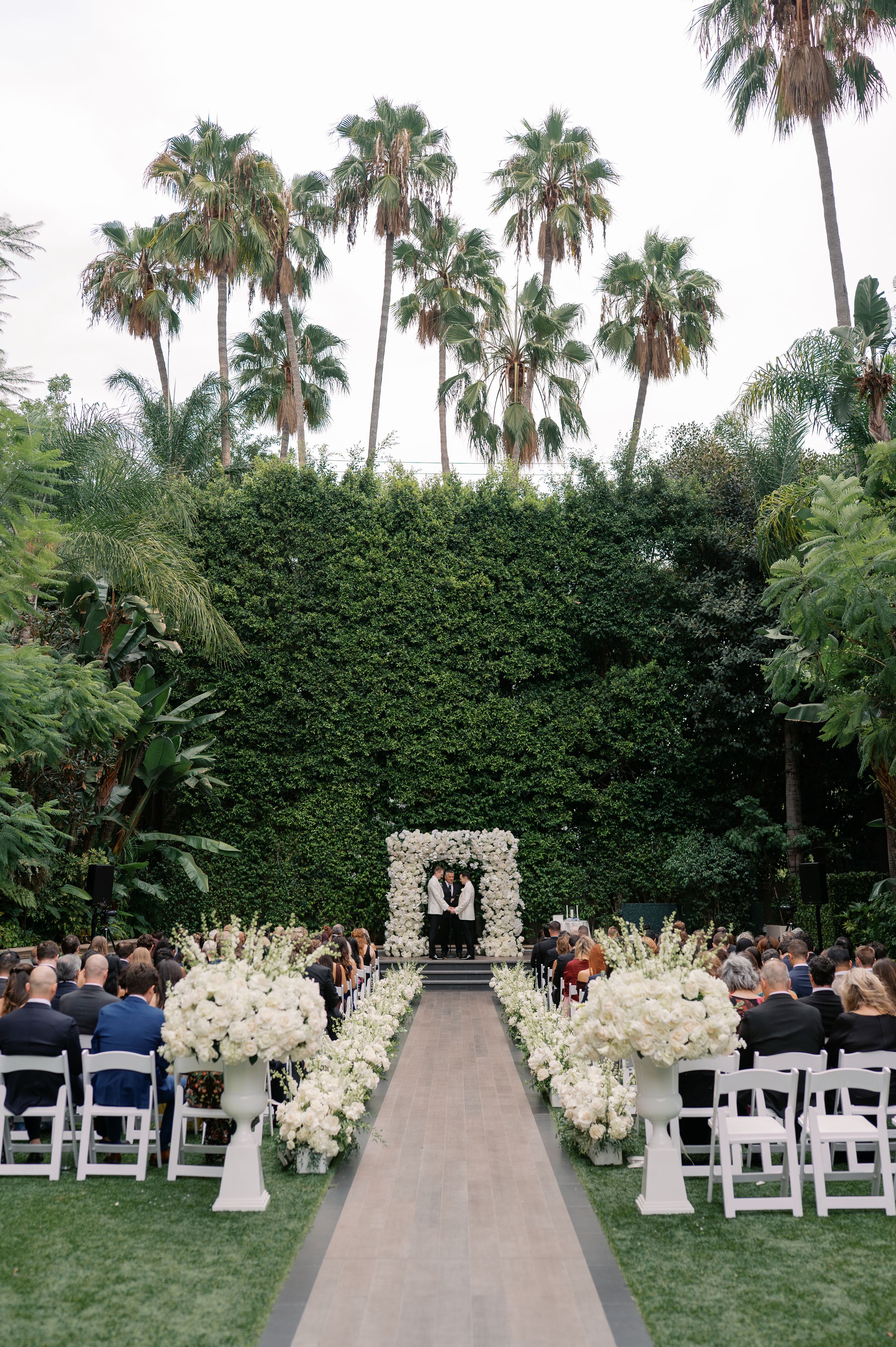 Elegant outdoor wedding ceremony with white floral arrangements lining the aisle, arch backdrop, and tall palm trees at a garden venue.