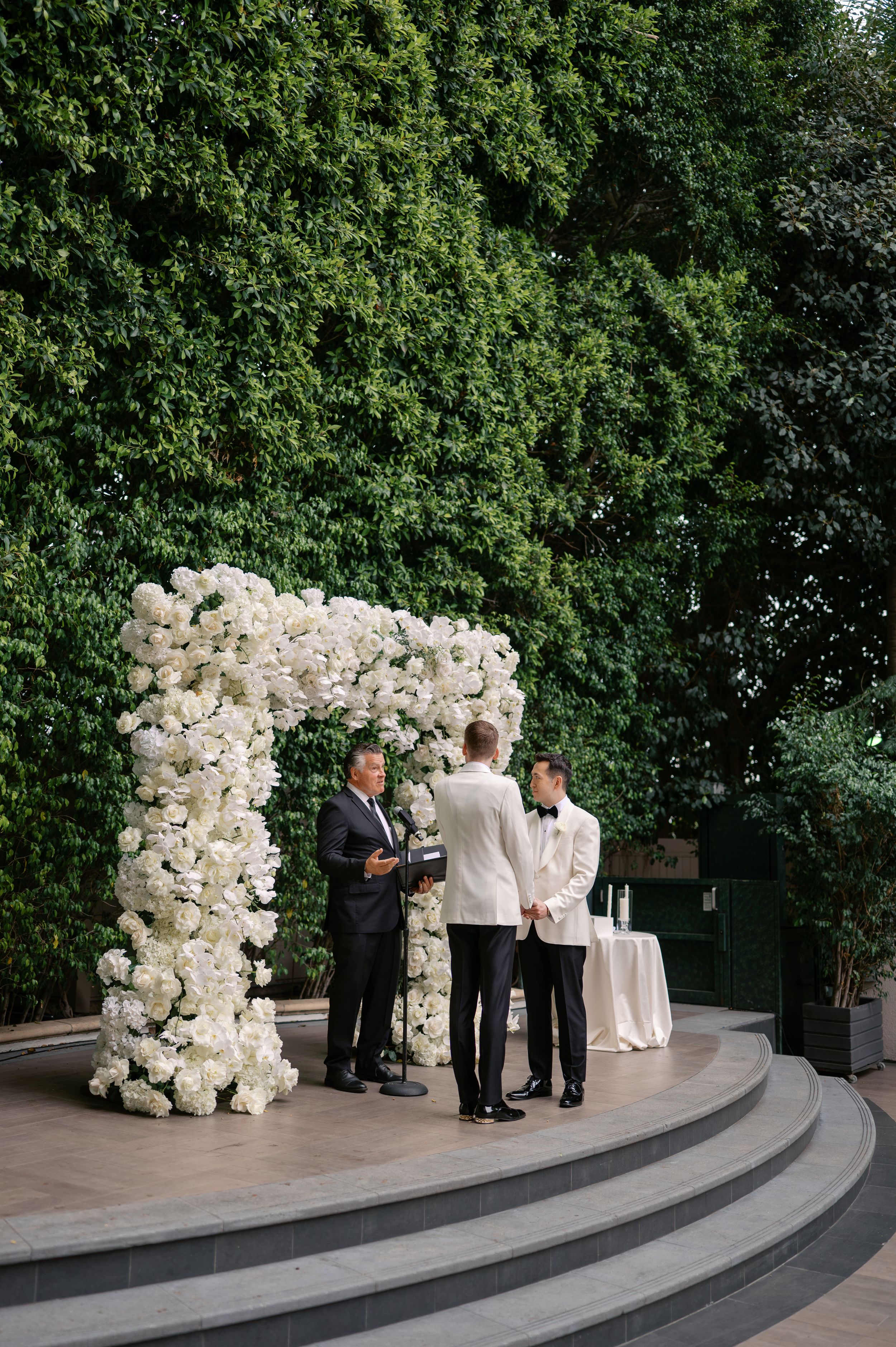 Two grooms exchange vows under a white floral arch at an elegant outdoor wedding ceremony.