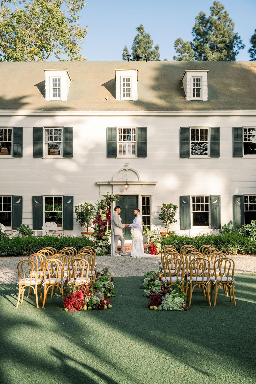 Outdoor wedding ceremony setup with wooden chairs facing white colonial house adorned with floral arrangements.