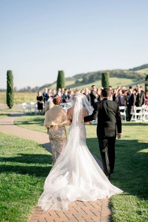 A romantic outdoor wedding scene with flowing white dress trailing behind on a grassy hilltop venue.