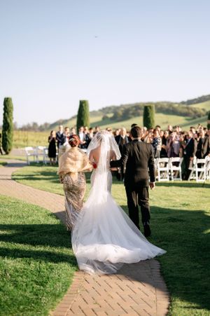 A wedding ceremony procession on a brick path with cypress trees and rolling hills in the background.
