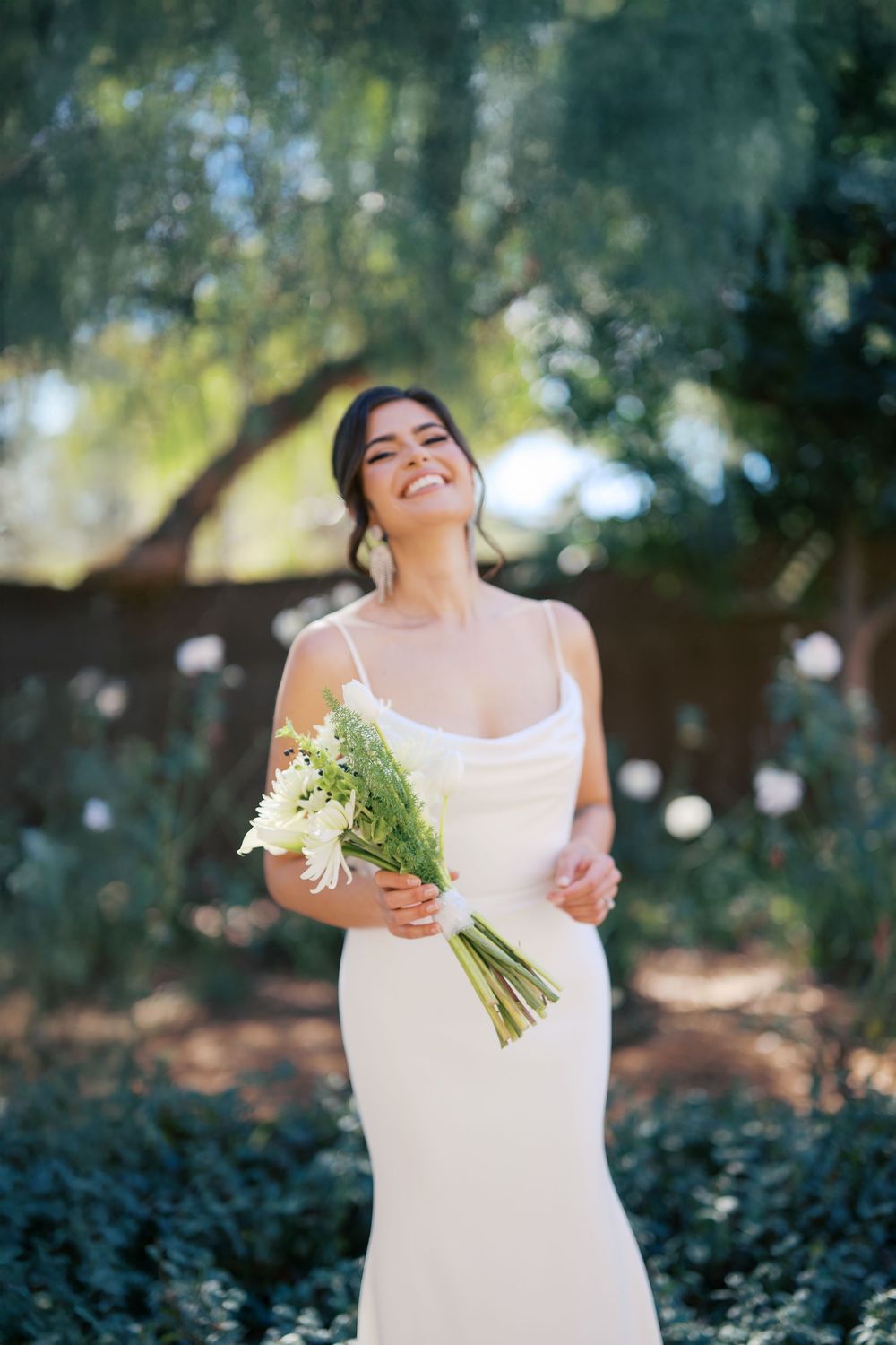 A joyful bride in a sleek white wedding dress holds a simple bouquet of daisies and greenery in a natural outdoor setting.