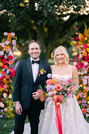Couple in black tuxedo and white wedding dress standing together with bright colorful floral arrangements at sunset.