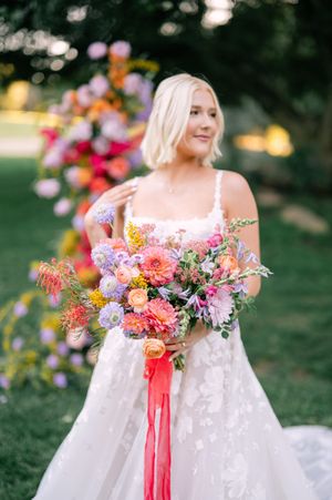 Bride in white lace gown holds vibrant summer bouquet with pink and coral flowers tied with flowing ribbon in garden setting.
