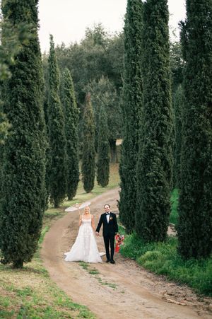 A couple walks down a cypress tree-lined path in elegant wedding attire.