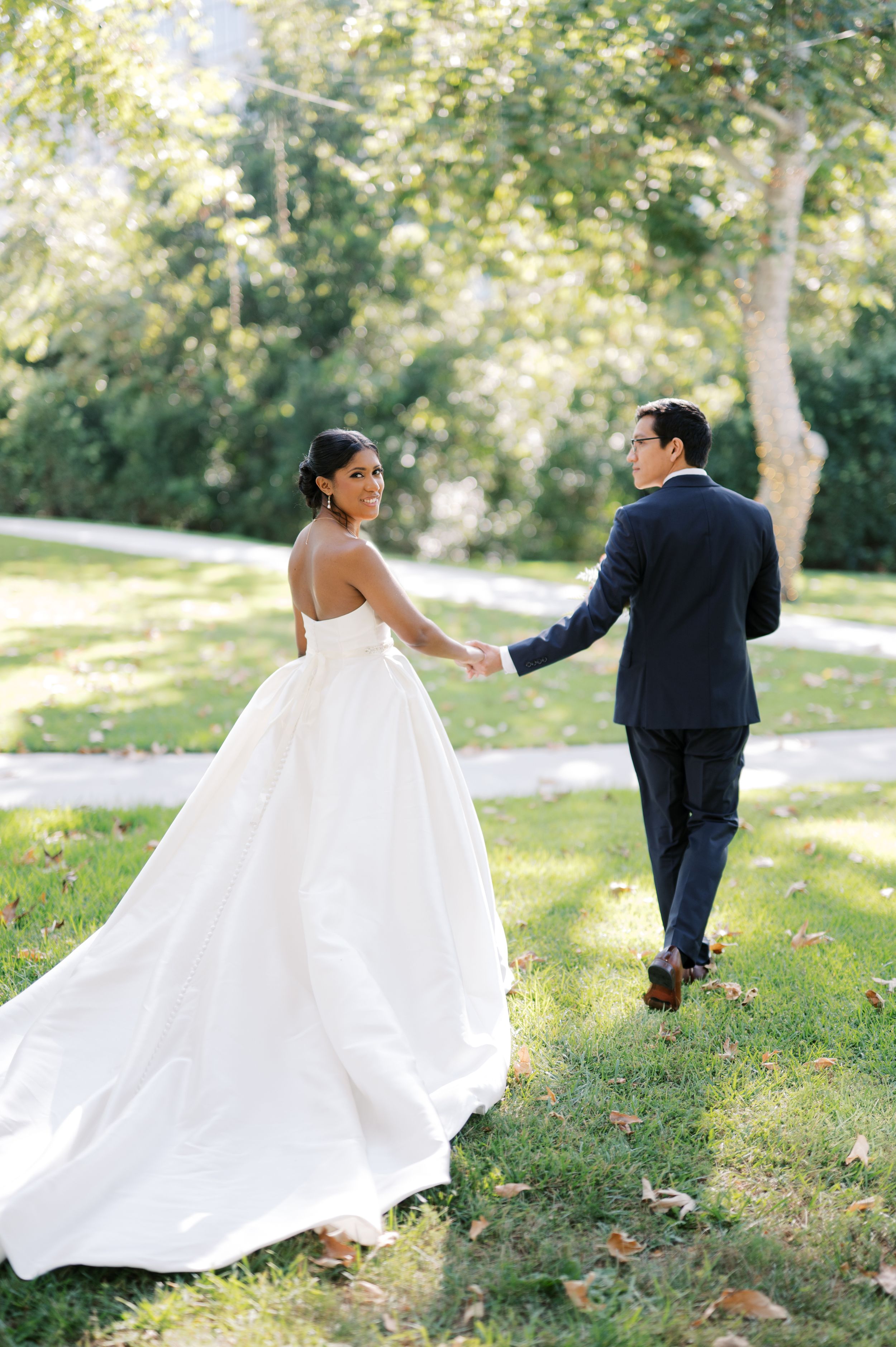A bride in a white ballgown and groom in navy suit walk together through a sun-dappled garden path.