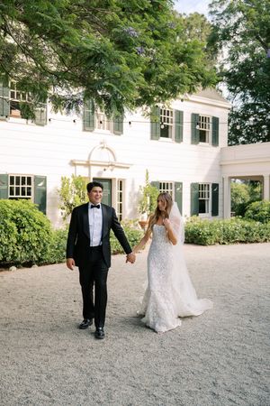 Bride and groom walk hand-in-hand outside a white colonial estate with green shutters on their wedding day.