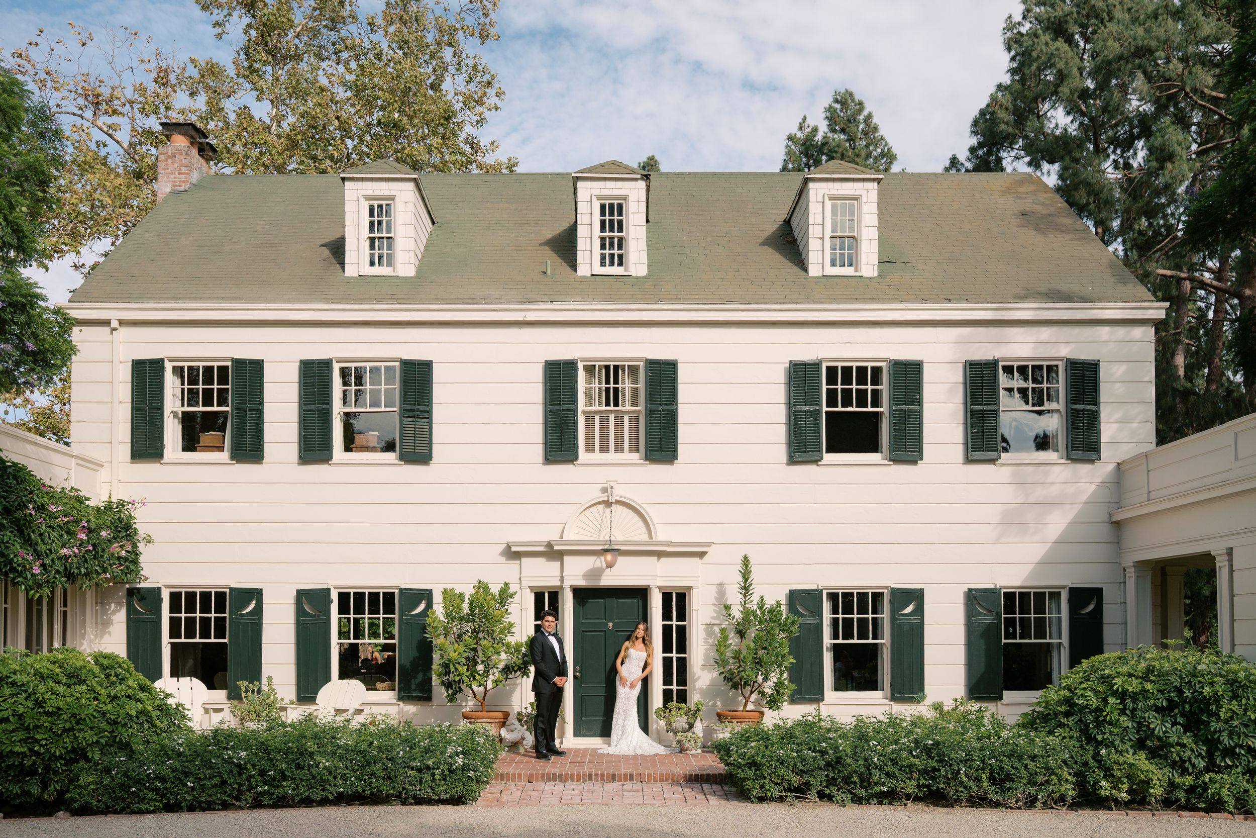 Bride and groom pose before a elegant white colonial wedding venue with dark green shutters and lush landscaping.