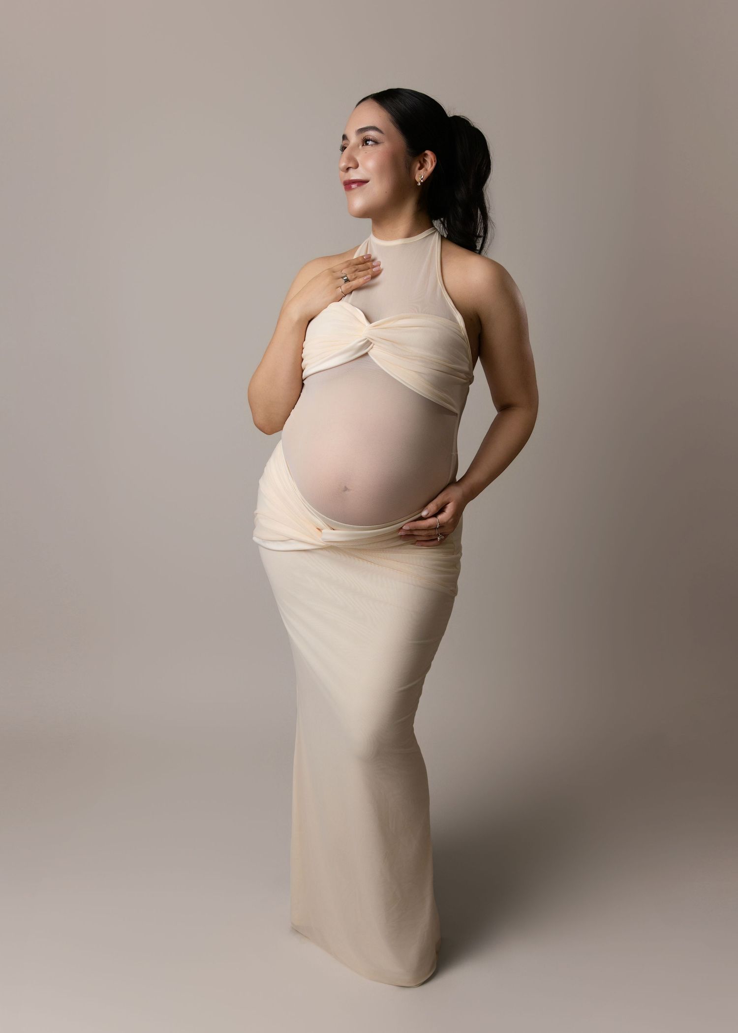 Pregnant woman in cream-colored fitted dress poses elegantly against neutral background during maternity photoshoot.