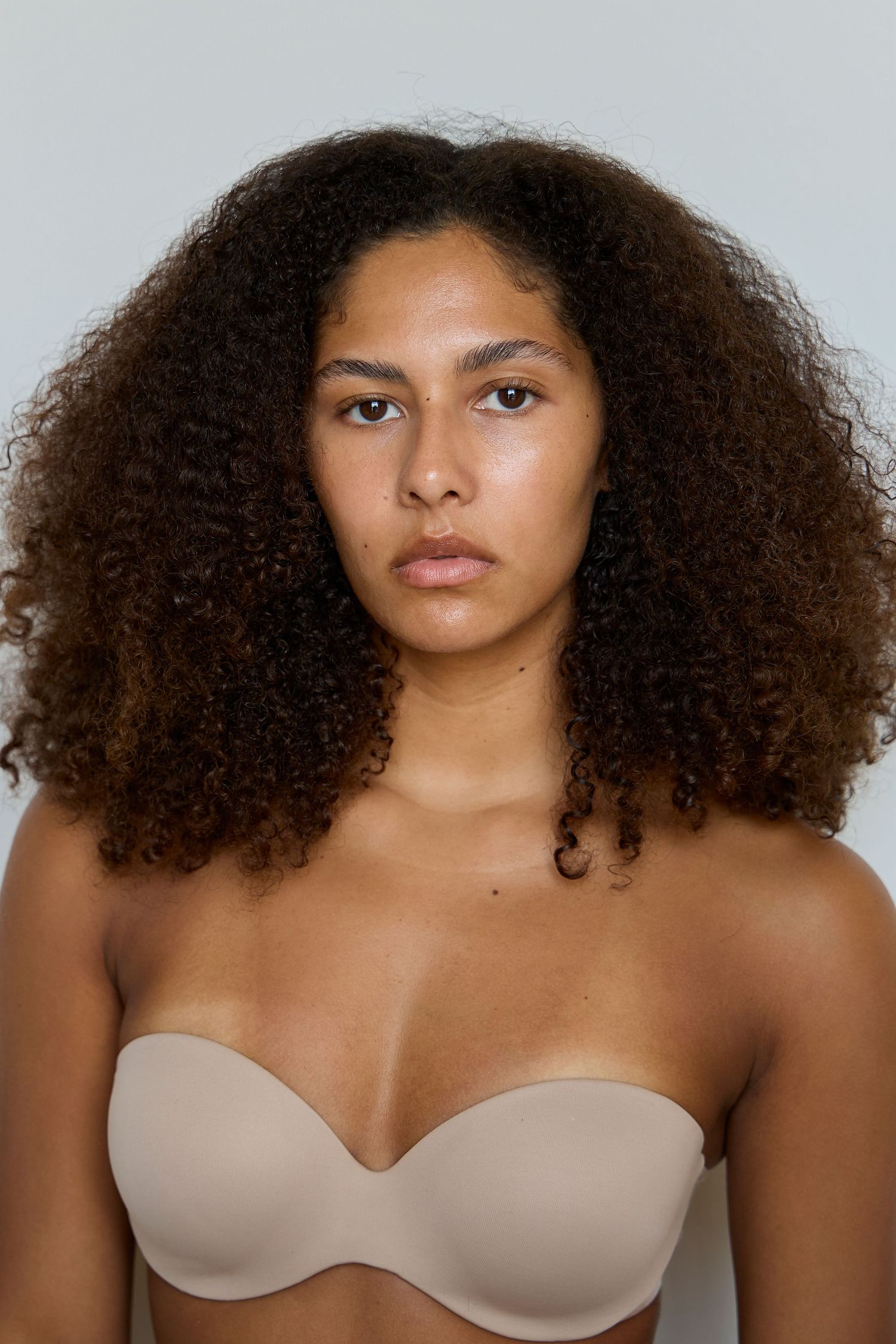 A woman with voluminous curly hair poses in a nude strapless bra against a neutral background.
