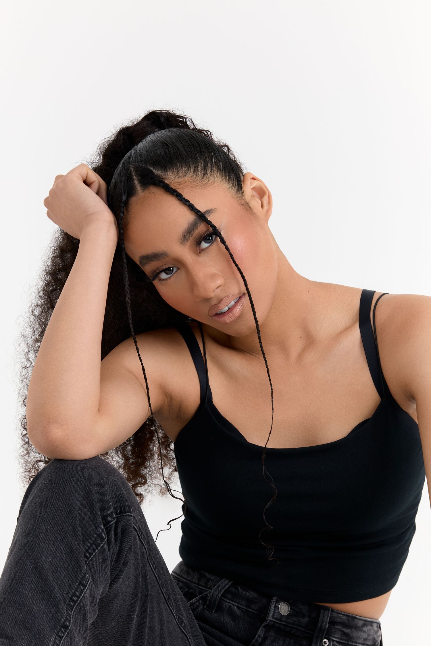 Fashion model in black tank top and jeans poses with curly hair against white background.
