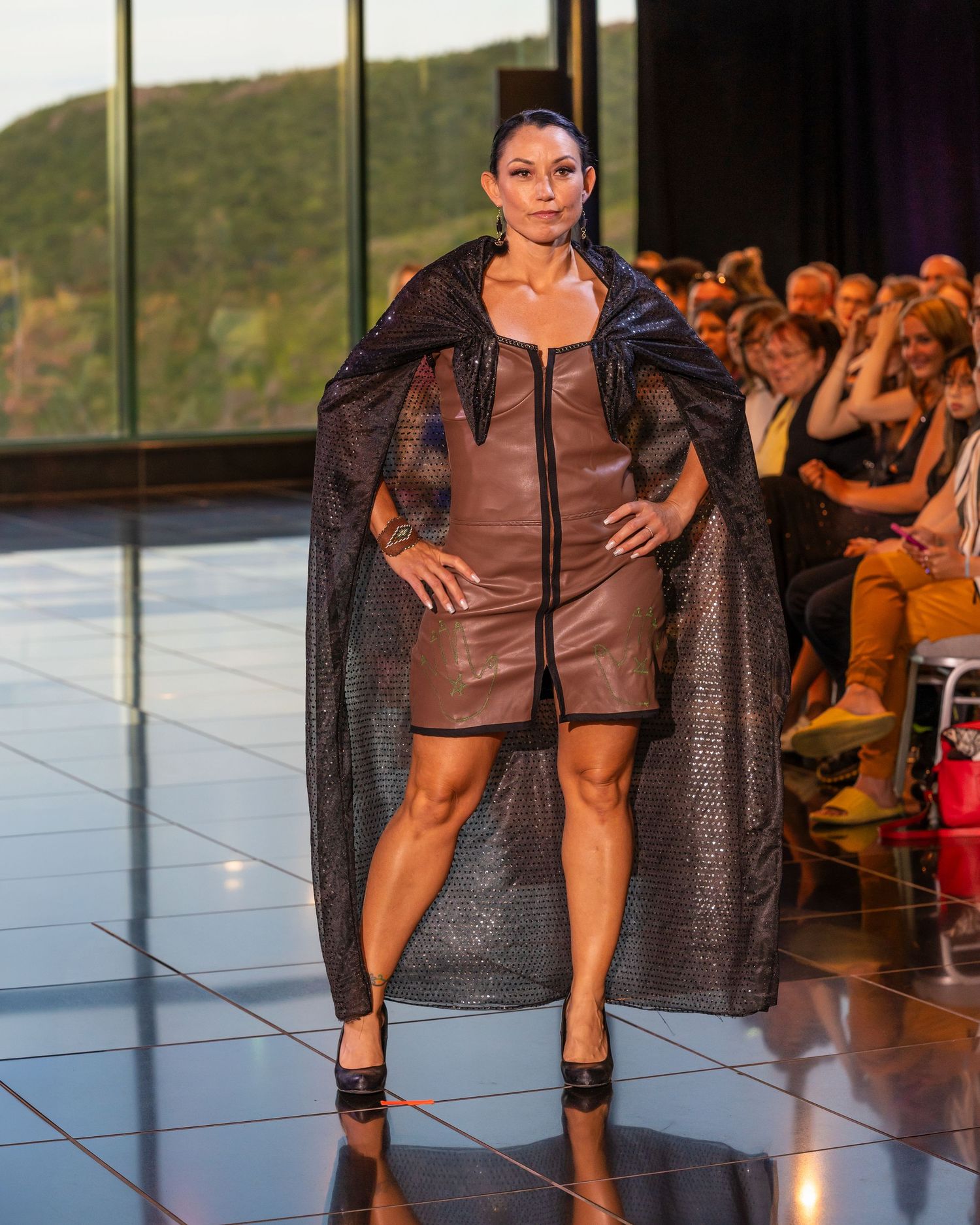 Model in brown leather dress and sheer black cape poses on runway with audience visible in background.