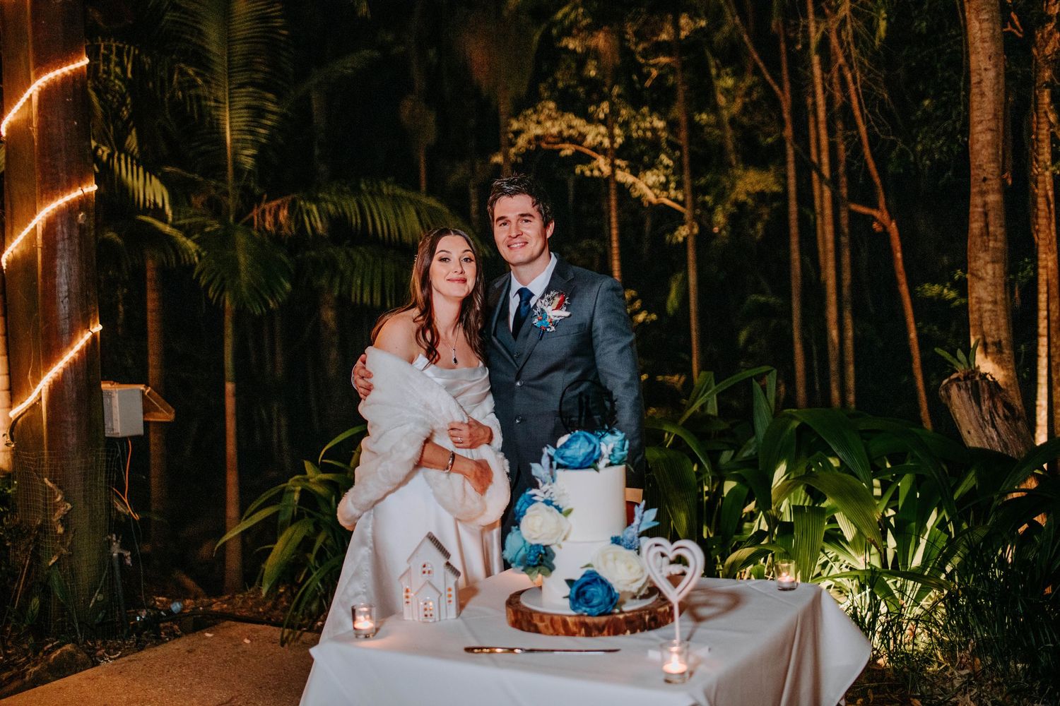 Newlyweds share moment while cutting their blue and white wedding cake at outdoor reception.