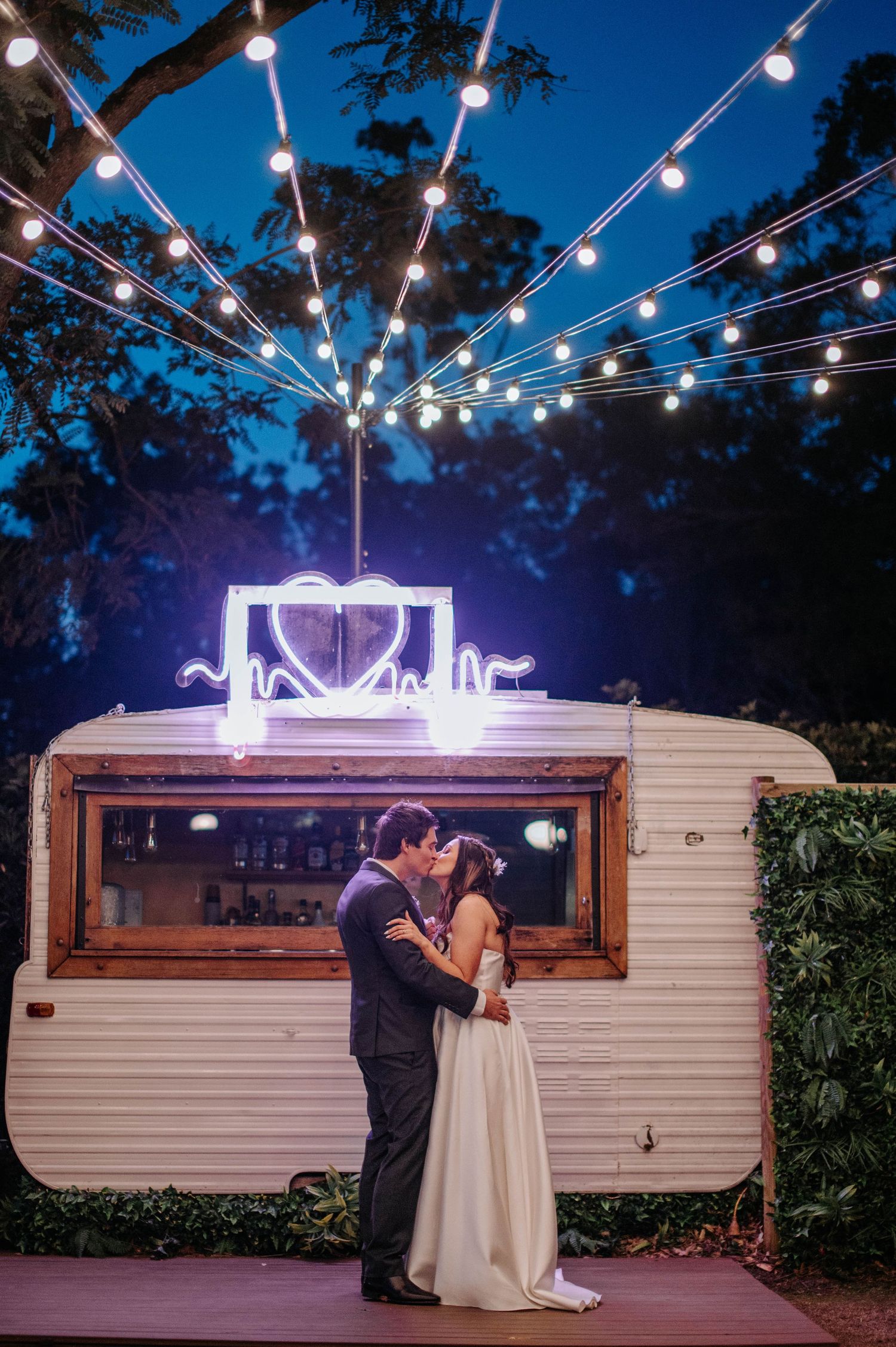 Romantic evening wedding scene with string lights over vintage caravan trailer and couple sharing intimate moment.