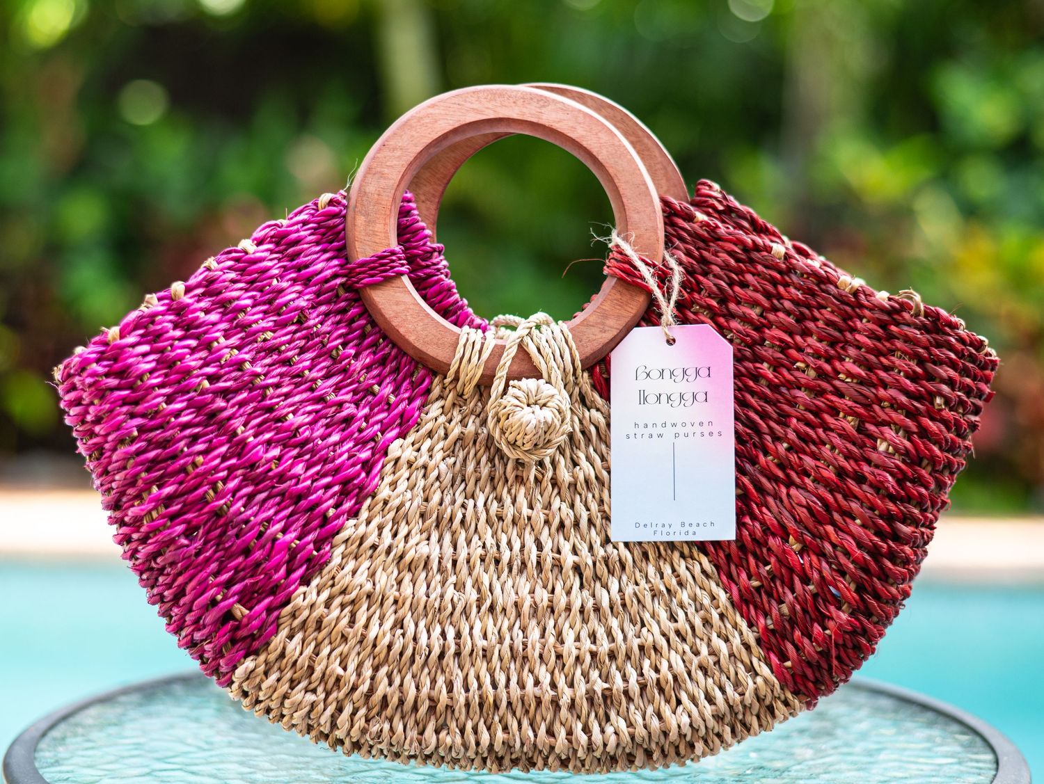 Pink and natural two-tone woven straw beach bag with wooden handles against blurred outdoor background.