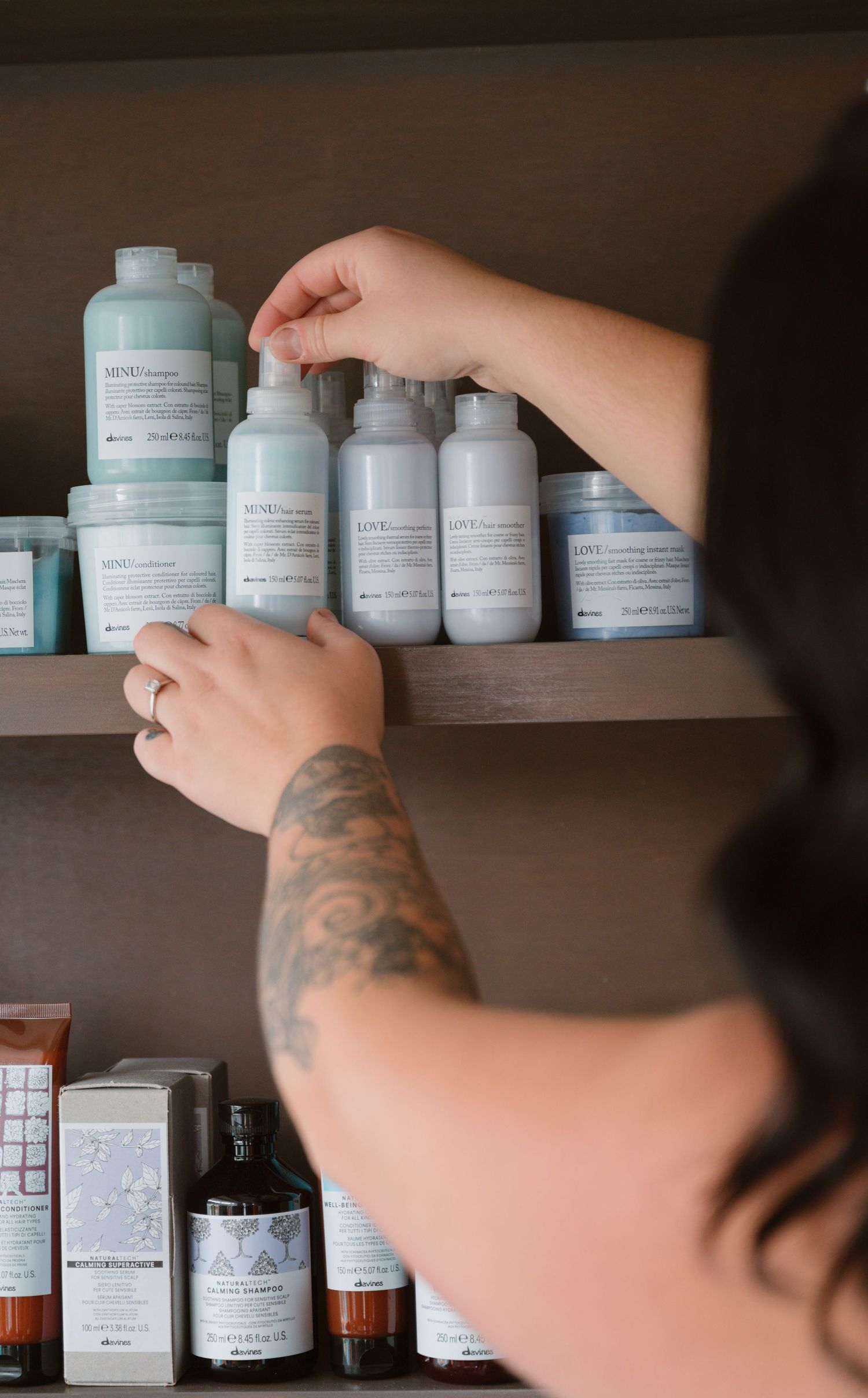 Tattooed hands organizing professional hair care products on salon shelf.