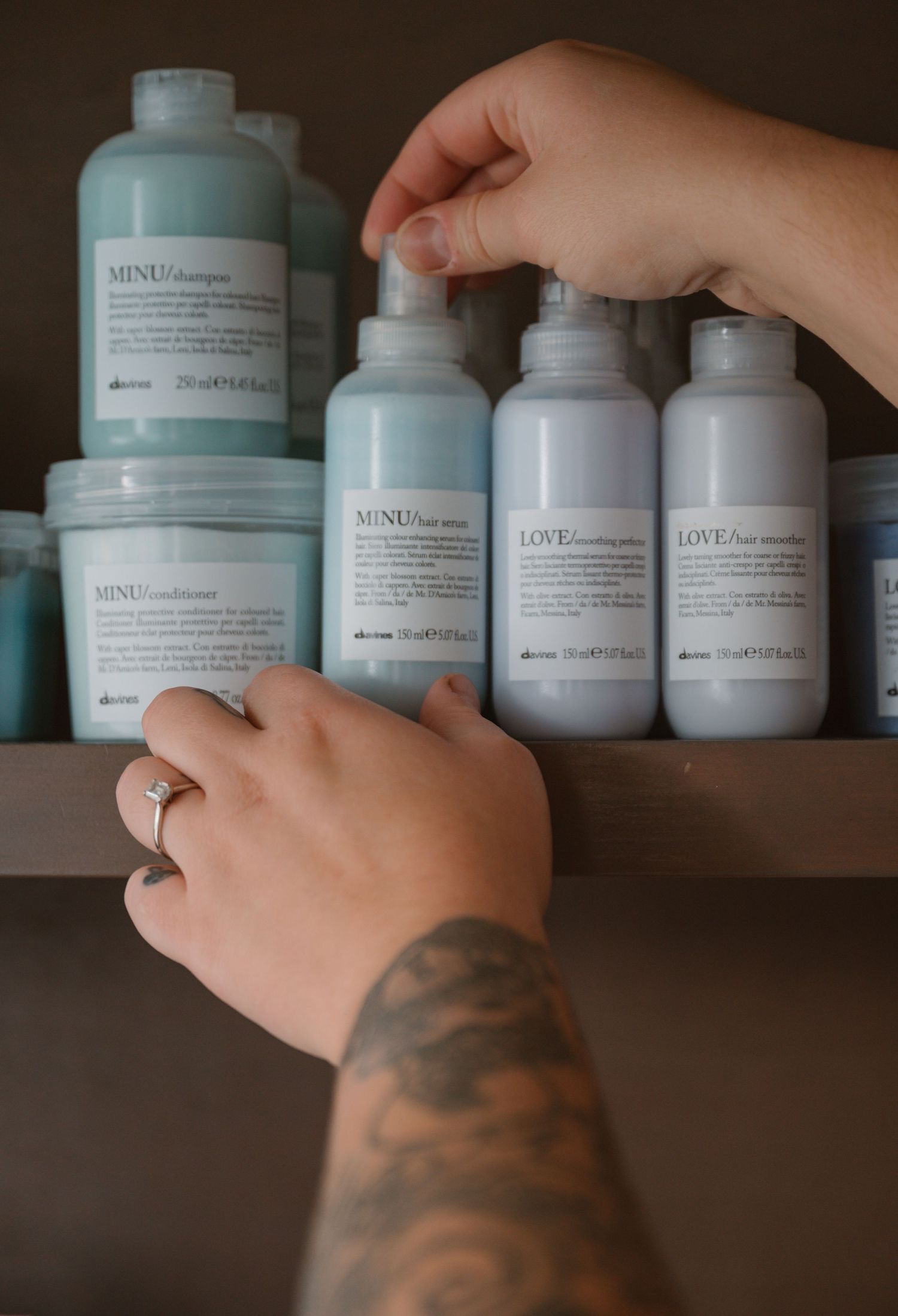 Hands with tattoos reaching for professional haircare products in mint green and white bottles on wooden shelf.