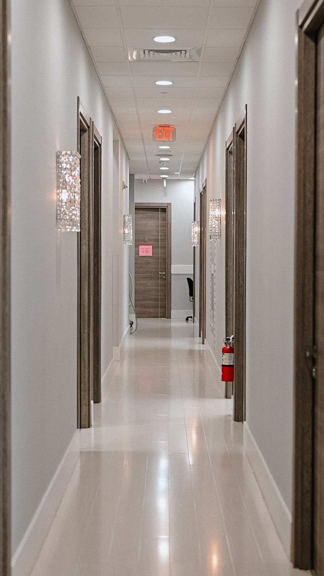 Modern office hallway with white walls, decorative sconces, and brown doors leading to an elevator.