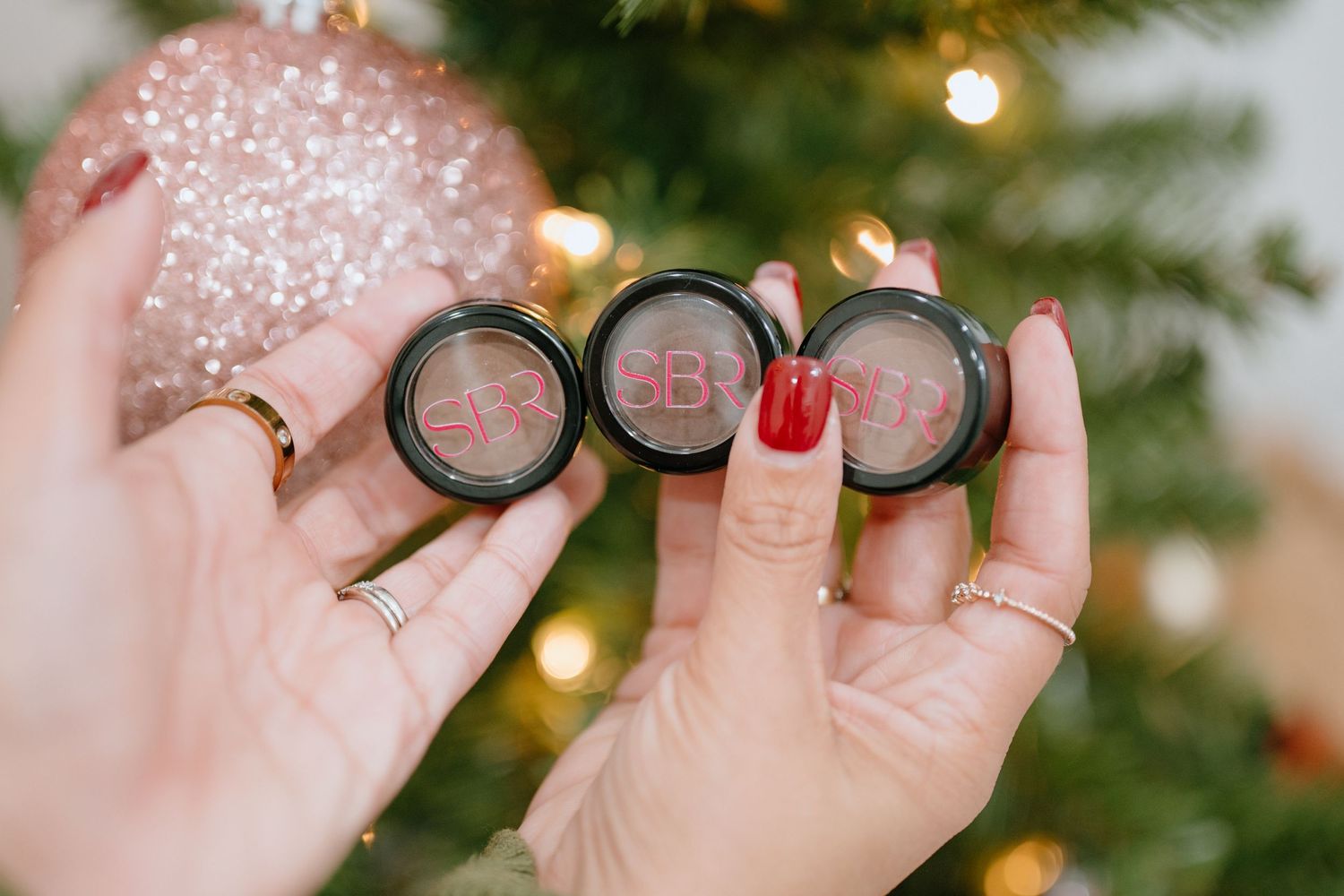 Hand holding three round makeup compacts against Christmas tree lights.
