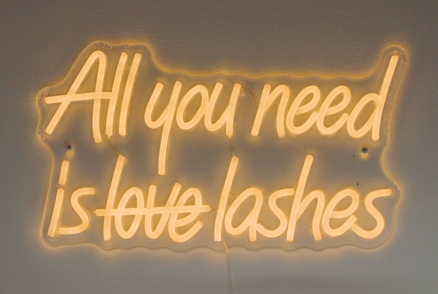 Yellow neon sign displaying 'All you need is lashes' against dark wall.