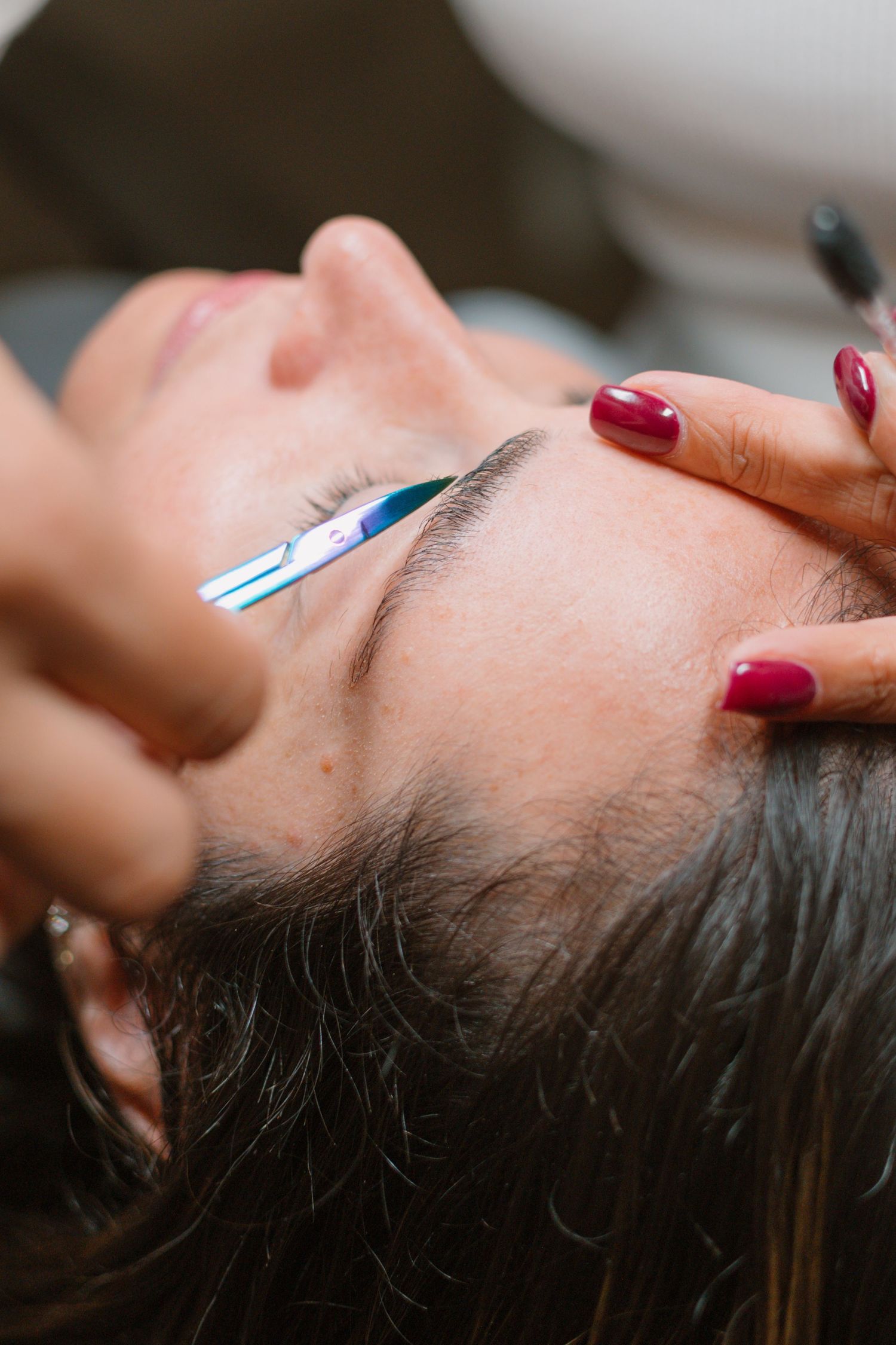 Close-up of eyebrow grooming procedure with tweezers and red manicured nails.