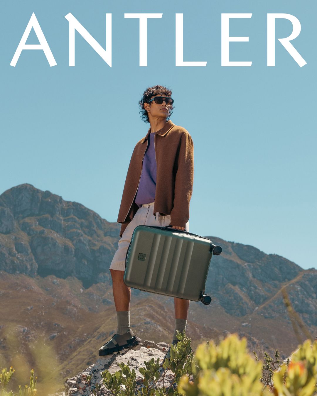 Person posing with grey hardshell luggage in mountainous terrain against blue sky.