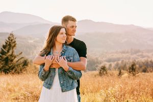A couple embraces in a scenic mountain meadow during sunset with golden grass and rolling hills in the background.