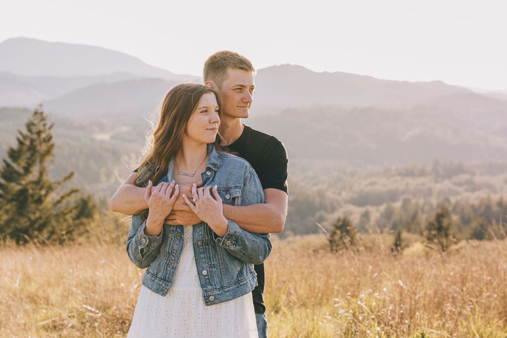 A couple embraces in a golden mountain meadow as they gaze at the scenic landscape during sunset.