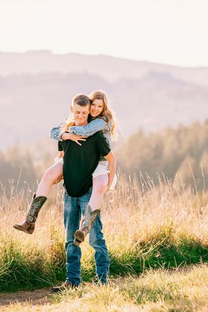 A couple embraces on a hilltop during golden hour with mountains and tall grass in the background.