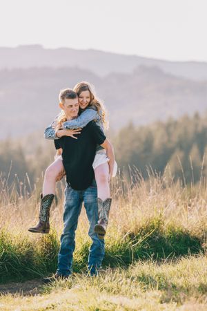 A couple embraces in a sunlit meadow with mountains and forest in the background during golden hour.