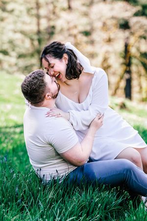 A romantic couple in white outfits lay together in soft green grass during a golden hour photoshoot.