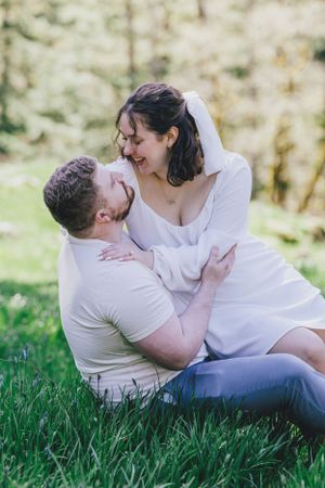 A couple in white clothing share a joyful intimate moment while sitting together on green grass in a sunlit outdoor setting.