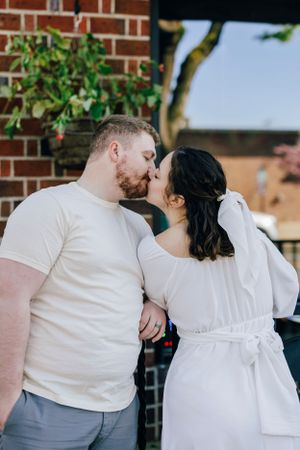 A couple in white outfits share a romantic kiss in front of a brick wall with green foliage.