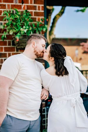 A romantic couple sharing a tender kiss against a brick wall backdrop with green foliage.