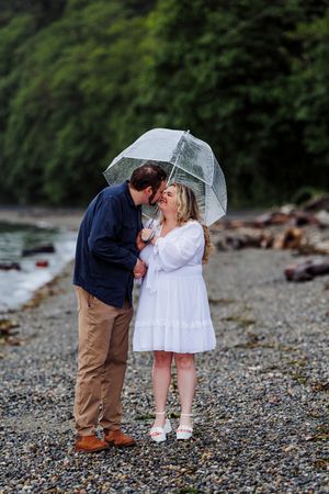 Couple shares romantic moment under clear umbrella on rocky beach shore with green foliage in background.