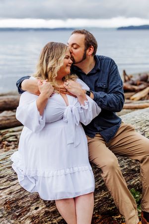Romantic couple embraces on driftwood at beach, with woman in white dress and man in navy jacket against ocean backdrop.