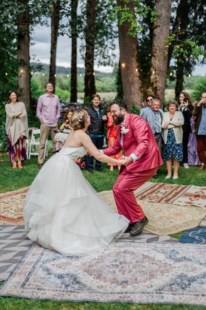 Wedding couple dances on vintage rugs laid outdoors with wedding guests watching under tall trees.