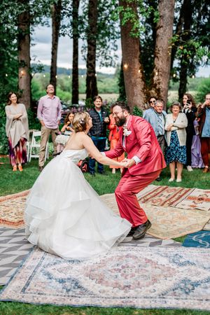 Wedding couple dances energetically on vintage rugs outdoors with trees and guests in background.