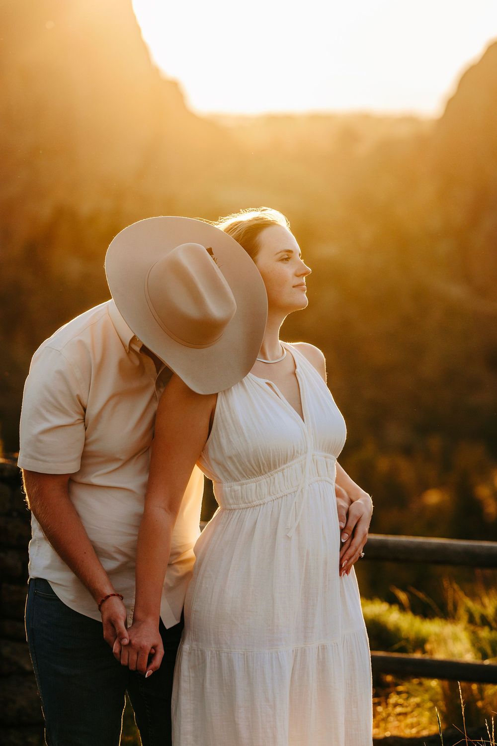 A couple in white attire embrace during golden hour sunset, creating a romantic silhouette against the warm glowing backdrop.