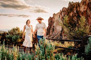 Couple walking hand in hand through desert landscape with tall cliffs and brush at sunset.