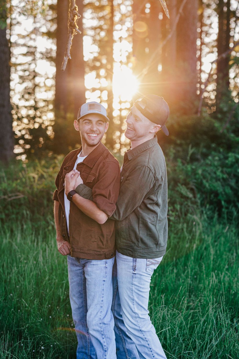Two people wearing casual clothing stand together in a grassy field at sunset with sun flare and trees in the background.